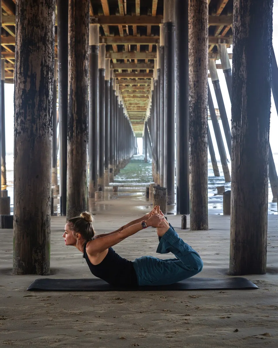 Anya Gossage in a yoga bow pose on a mat under a beach pier