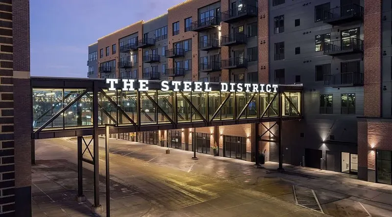 The Steel District exterior at dusk &mdash; illuminated steel-bridge signage spanning between brick buildings