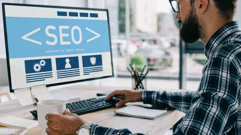 A man in a plaid shirt works at a desk, viewing an SEO interface on a computer monitor, researching the advantages of choosing a local SEO company for his business.