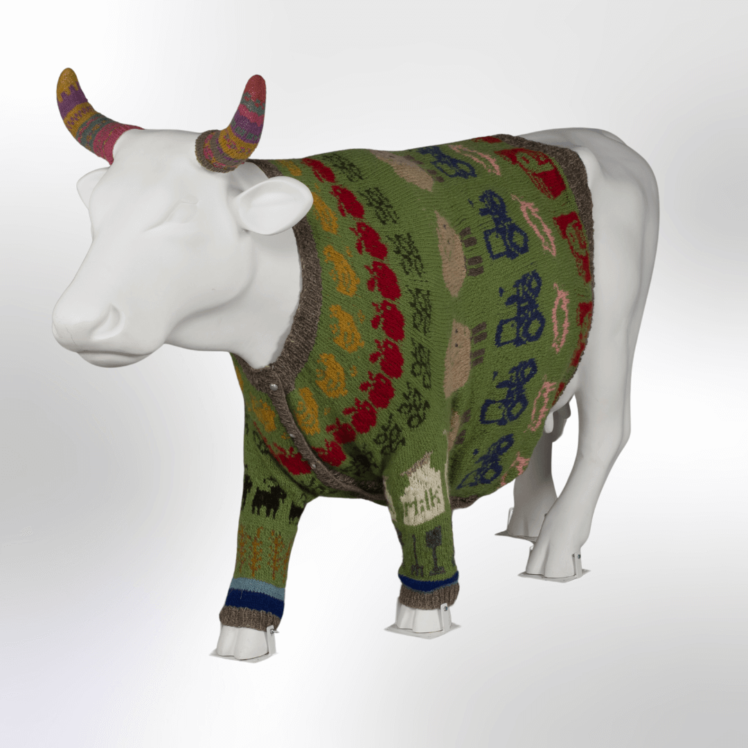 A cow wearing a hand-knit sweater featuring chickens, apples, sheep, tractors, and other farm motifs. The cow is also wearing horn warmers. 