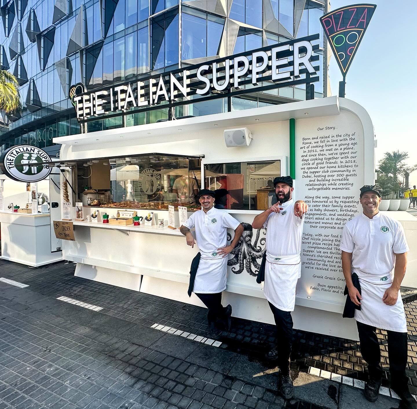 Chef at The Italian Supper food truck in Dubai smiling beside fresh garlic, olive oil, and the restaurant’s wooden menu stand.