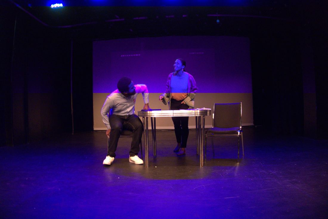 Two actors at a table under stage light