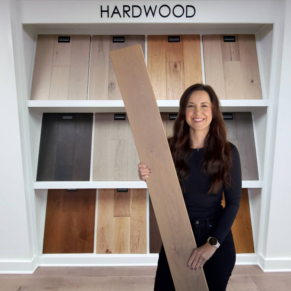 Jenna with AZ Floor Haven in southern Arizona holding a hardwood flooring sample in front of a display featuring various wood flooring options, labeled "HARDWOOD," illustrating personalized flooring selection for home design.
