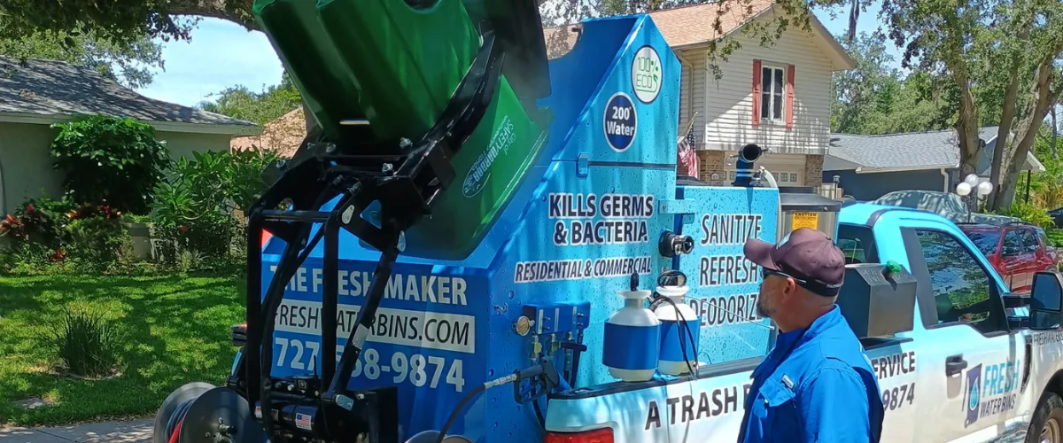 A blue sanitation truck with a man operating a mechanism cleaning a green trash bin on a sunny residential street. A blue sanitation truck with a man operating a mechanism cleaning a green trash bin on a sunny residential street.