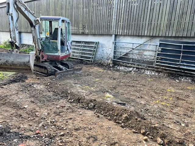 Mini digger excavating a yard beside a farm building