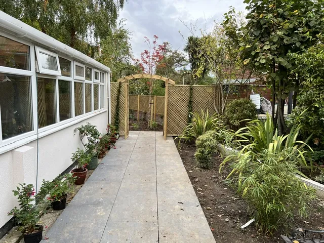 Paved path between planted beds beside a white-rendered bungalow
