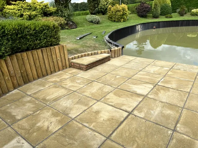 Buff paved patio with log-roll edging beside a garden pond