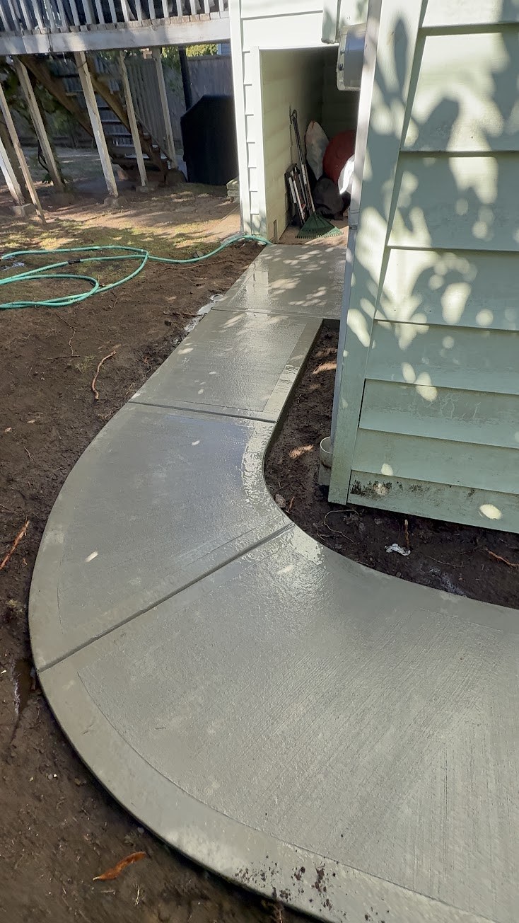 Curved concrete walkway wrapping around the back corner of a house, wet surface with a garden hose nearby in South Vancouver