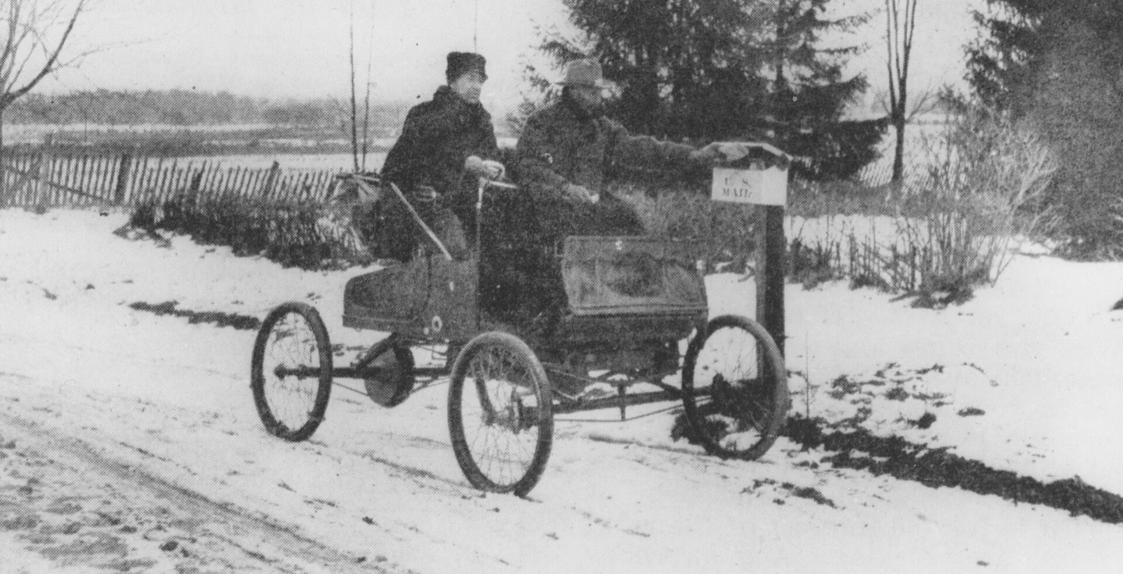 A rural mail carrier in an early automobile reaching into a roadside U.S. Mail box in the snow
