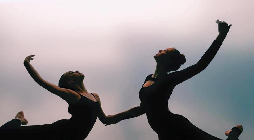 Two dancers in black silhouetted against a soft pastel sky, arms extended