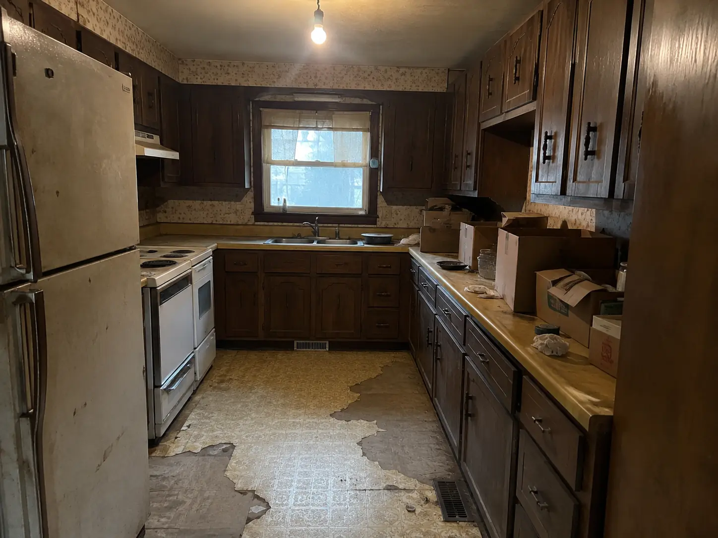 Dated 1960s kitchen with dark cabinets, torn vinyl floor and boxes on the counters