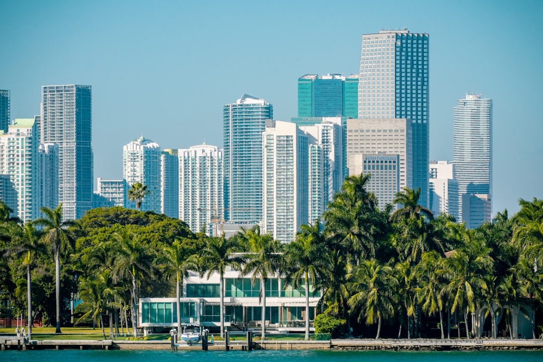 Miami downtown high-rise skyline behind a palm-lined waterfront with a low glass house at the water's edge.