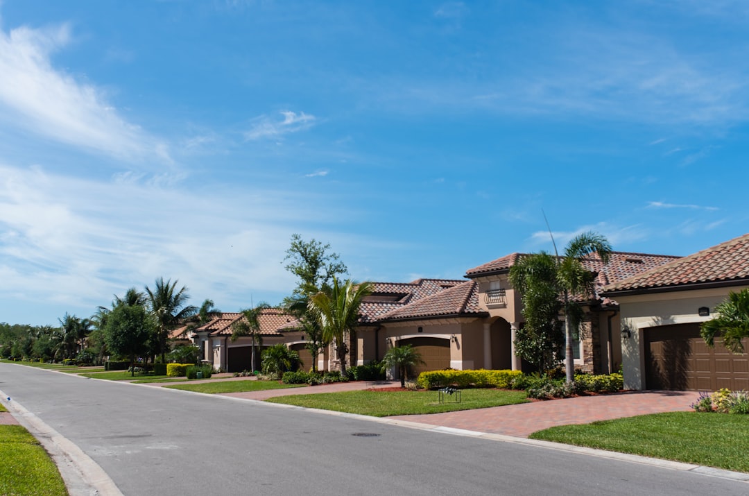Empty suburban street lined with tile-roofed stucco homes, palms and clipped lawns under a blue sky.