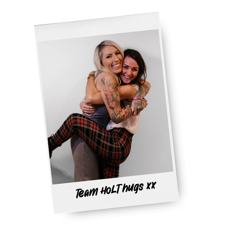 Two women embracing and smiling, one giving the other a piggyback ride, with tattoos on her arms, in front of a plain white background, with the caption "Team HOLThugs XX" at the bottom.