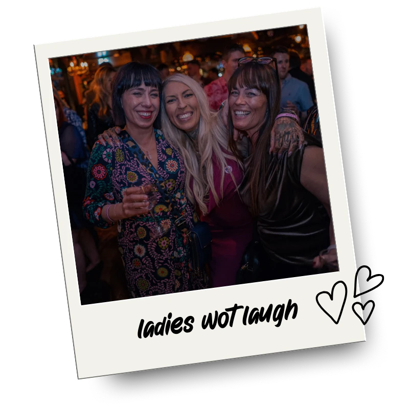 Three women smiling and laughing together at a social event or party, with a lively background and warm lighting.