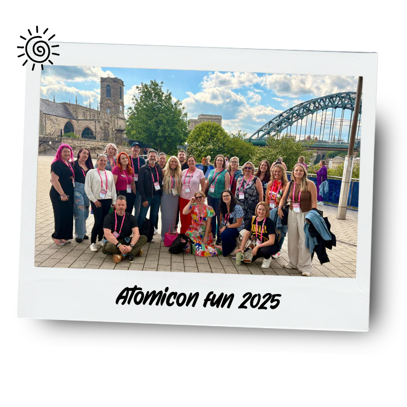 Group of people posing outdoors near a river with a bridge and historic building in the background, during daytime. The photo is framed like a polaroid with handwritten-style label "Atomicon fun 2025" at the bottom.