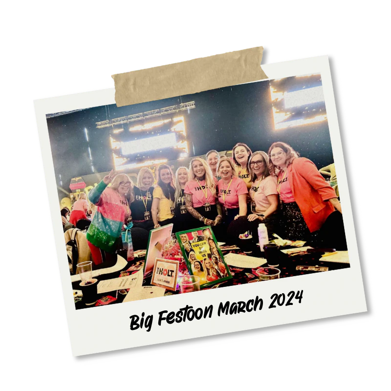 A group of women smiling and posing at a conference or event, with a table in front of them displaying various items and colourful posters. The background shows a stage with bright lights. The photo is framed as a polaroid with tape at the top and the caption "Big Festoon March 2024" written at the bottom.