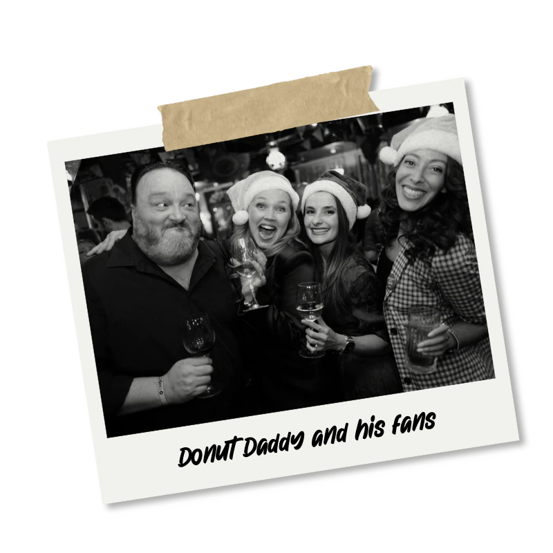 Group of four people celebrating with drinks, wearing Santa hats, at a holiday party, in black and white, with the caption 'Donut Daddy and his fans'.