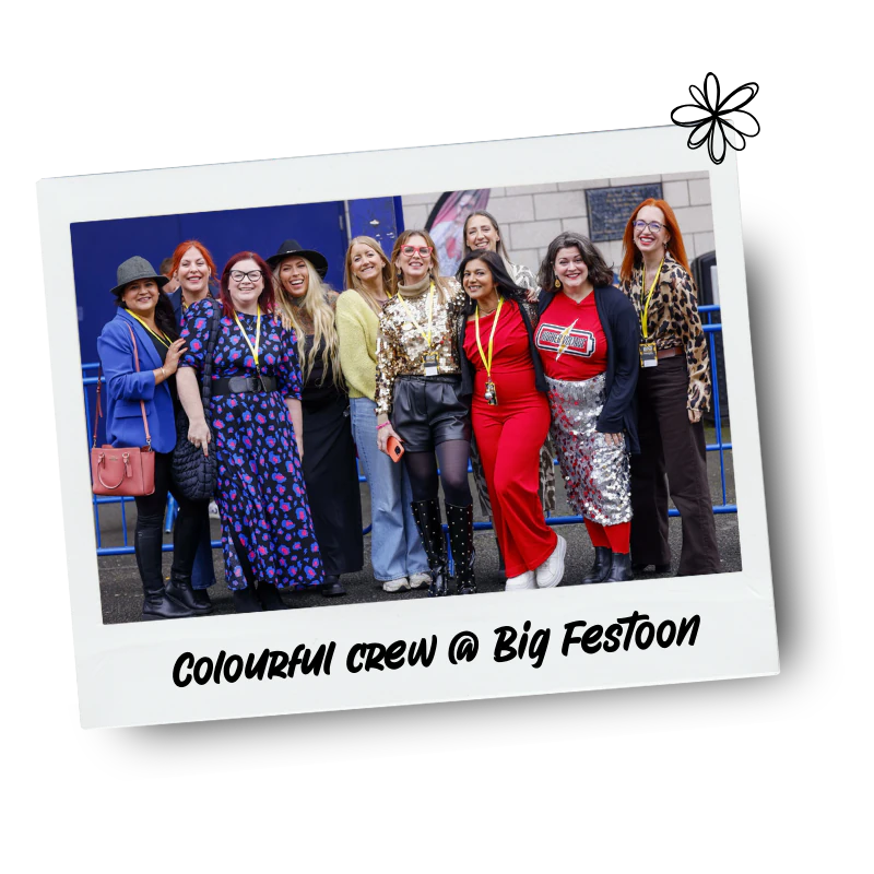 Group of women standing together outdoors, dressed in colourful outfits, smiling for a photo at Big Festoon.