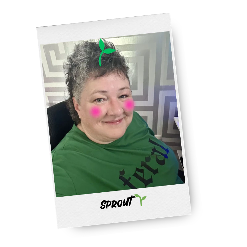 A woman with short, curly grey hair, wearing a green shirt with black text, sitting in a black chair, smiling. She has pink cheeks and a small green sprout graphic edited near her head.