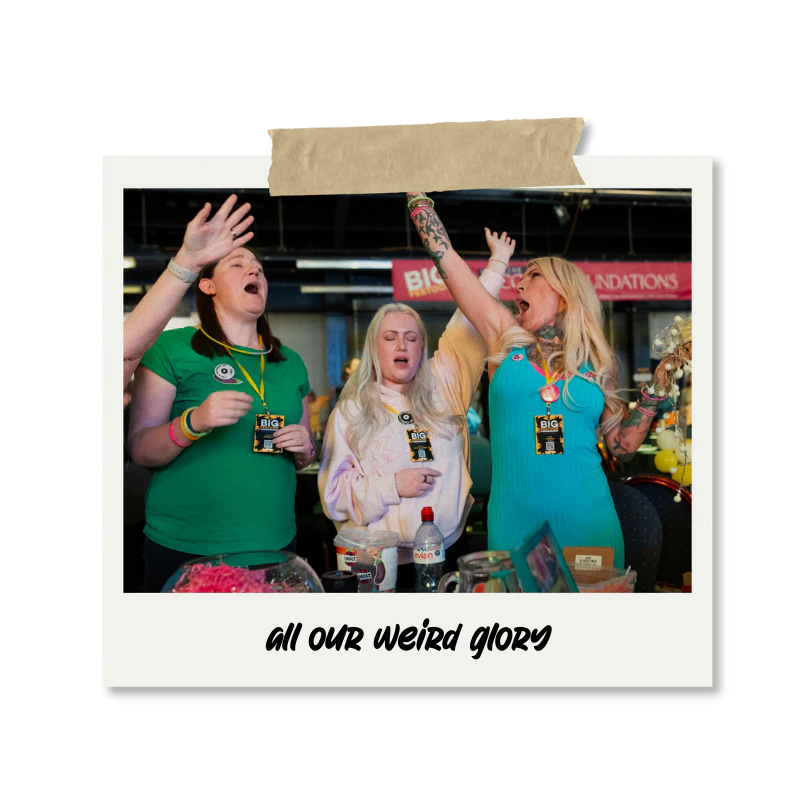 Three women at a celebration or convention, with one woman raising her arm and another closing her eyes, displaying a moment of enthusiasm or emotion, surrounded by food and drinks, with a banner in the background that says 'BIG'.