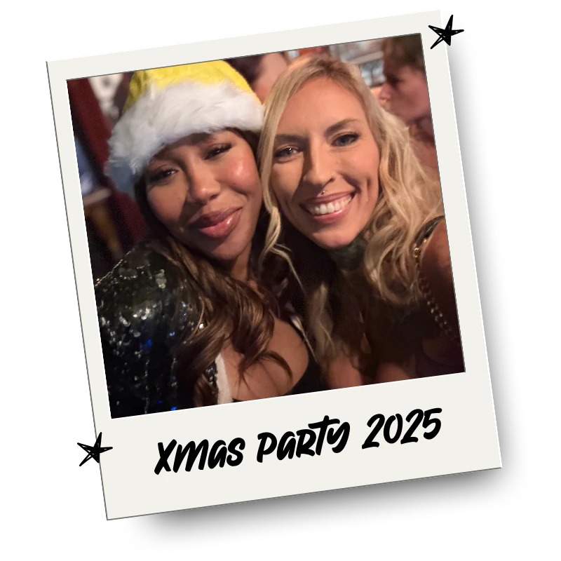 Two women smiling and taking a selfie at a Christmas party, one wearing a Santa hat, with a festive background.