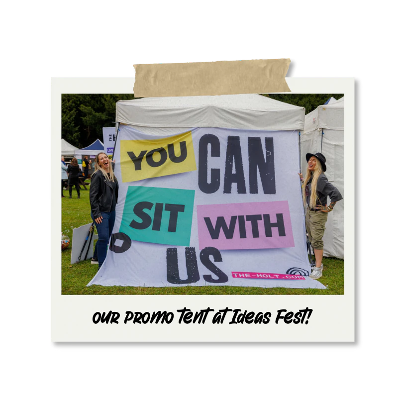 Two women standing in front of a promotional tent at Ideas Fest, with a sign that reads 'You Can Sit With Us,' in colourful block letters. One woman is smiling and wearing a leather jacket, while the other is laughing and wearing a hat and khaki pants.