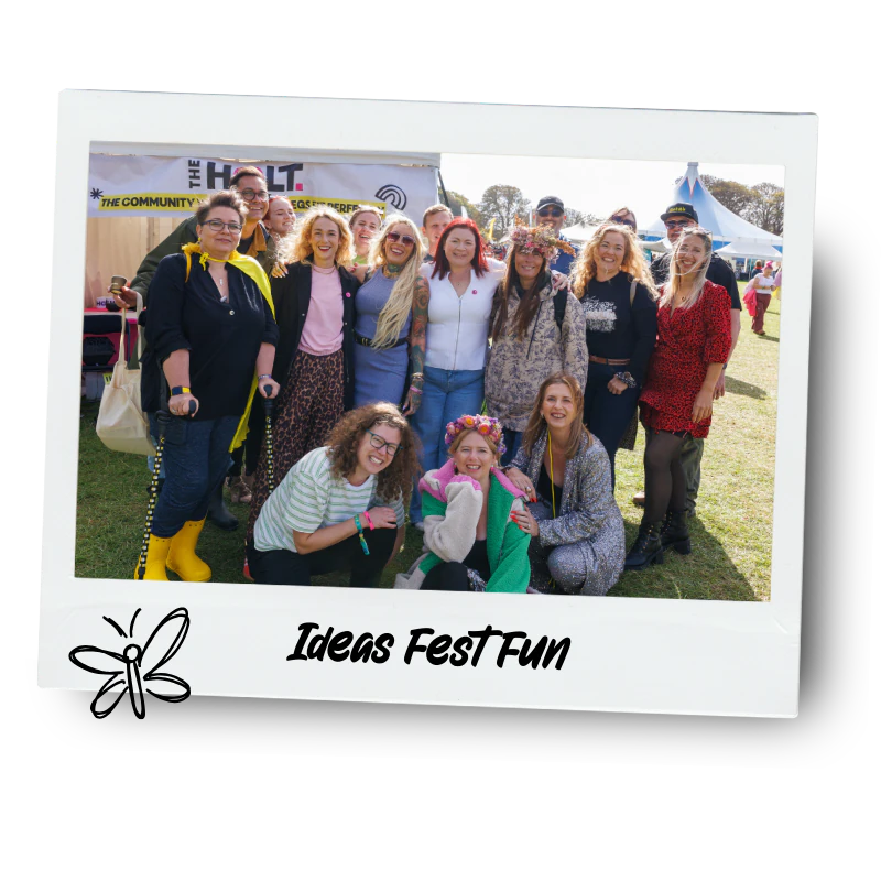 Group of people at Ideas Fest Fun event, outdoors with tents and banners in background, some wearing colourful or festive outfits, smiling for the camera.