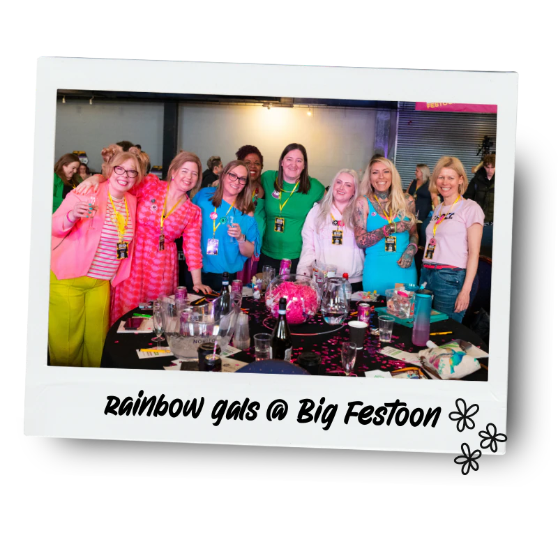 Group of women smiling and posing around a table at a big celebration event. They are wearing colourful outfits and lanyards. The table has drinks and decorations. Caption reads "rainbow gals @ Big Festoon" with flower icons.