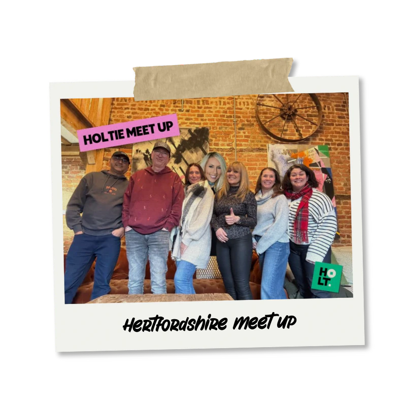 A group of seven people posing together inside a brick-walled room with artwork and a wagon wheel decoration, with a label at the top that reads 'HOLTIE MEET UP' and a caption at the bottom that says 'Hertfordshire meet up.'