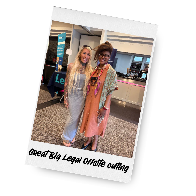 Two women standing together smiling at an indoor event, with a bar or counter in the background. One woman has long blonde hair and is wearing a light grey dress, and the other woman has curly hair with glasses and is dressed in an orange dress with accessories.