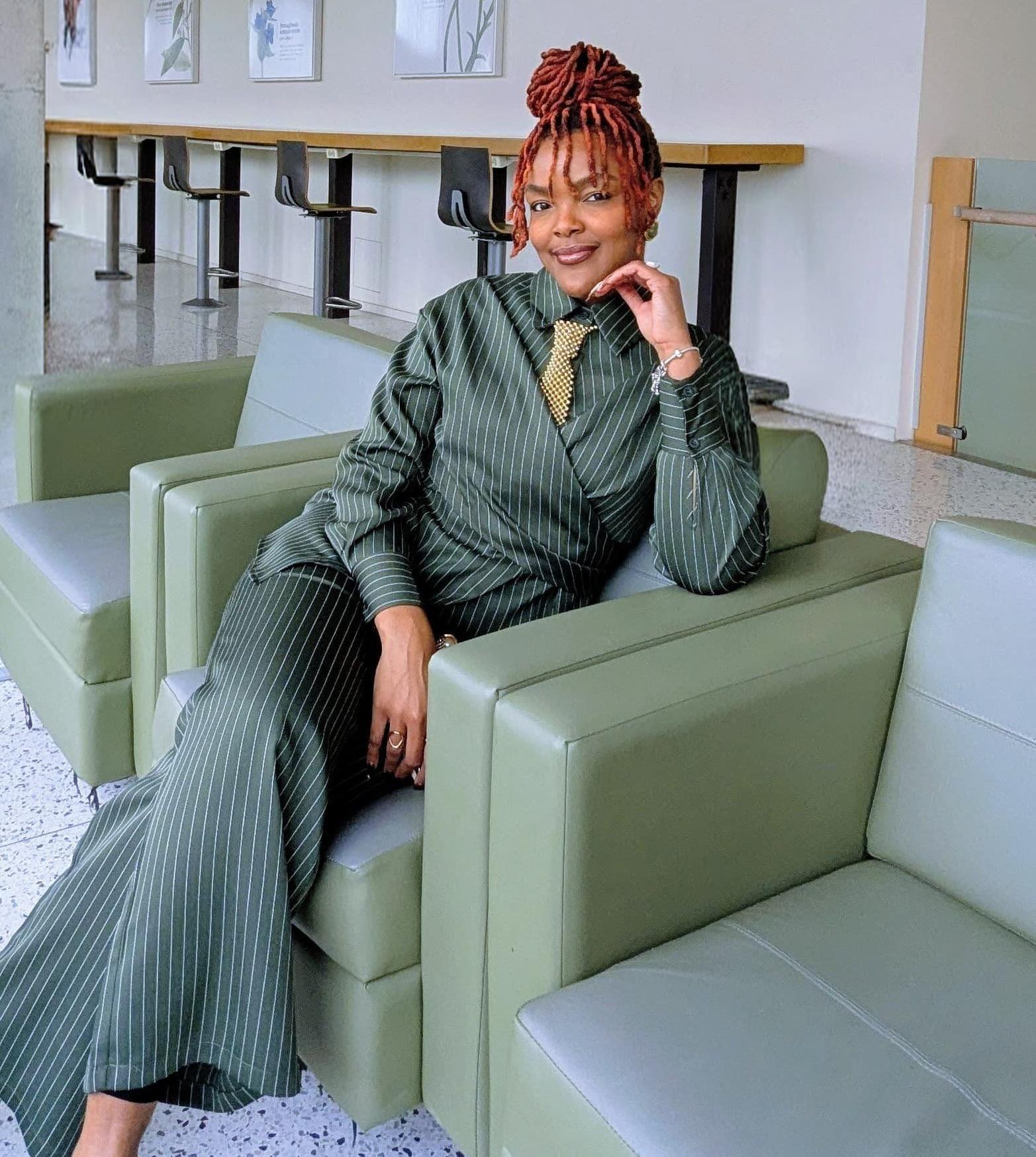 A stylish person with red braided hair wearing a green pinstripe suit and gold tie, seated confidently on a modern light-green lounge chair in a bright contemporary interior.