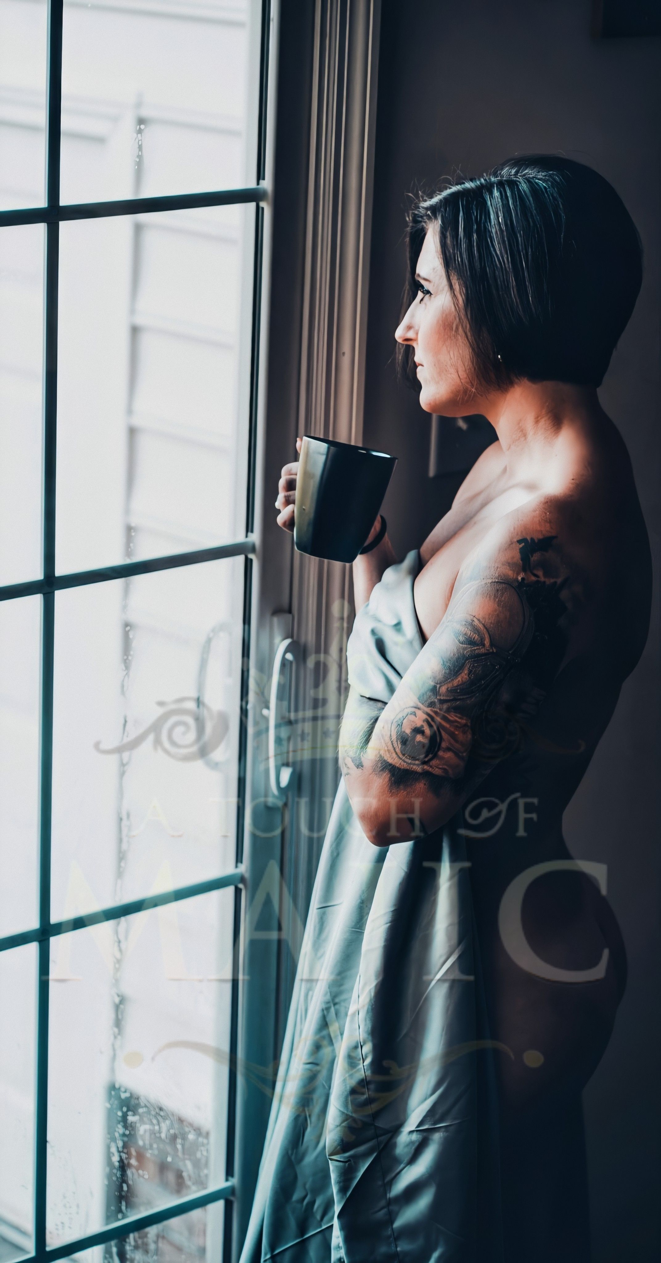 Sensual thirty something woman seductively wrapped in a sheet drinking coffee while standing next to window in Boudior pose