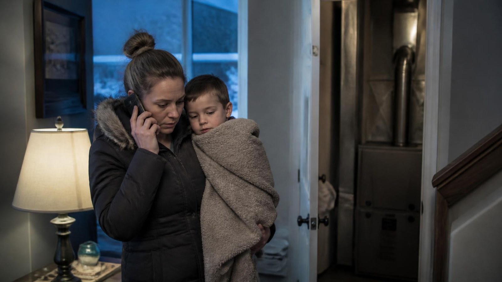 Cold mom calling an HVAC company for heating help A mom in a winter coat holds her bundled-up son and calls for heating repair next to an open furnace closet.