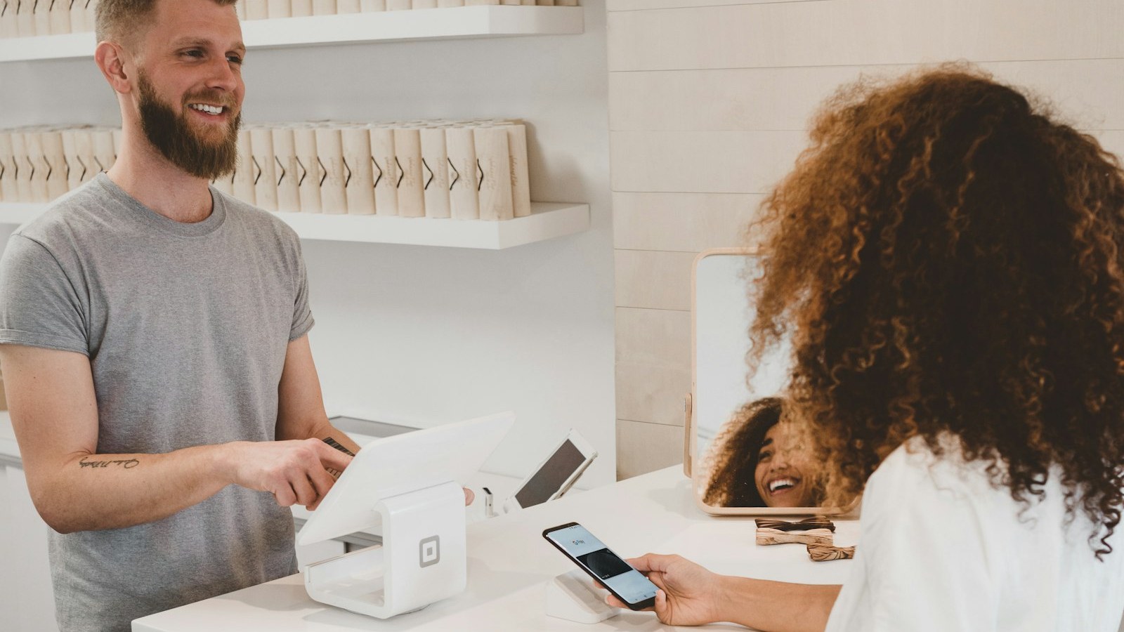Boutique counter handoff: staff at a tablet POS helping a customer with a phone — pickup and repeated-question service cue. Boutique counter handoff: staff at a tablet POS helping a customer with a phone — pickup and repeated-question service cue.