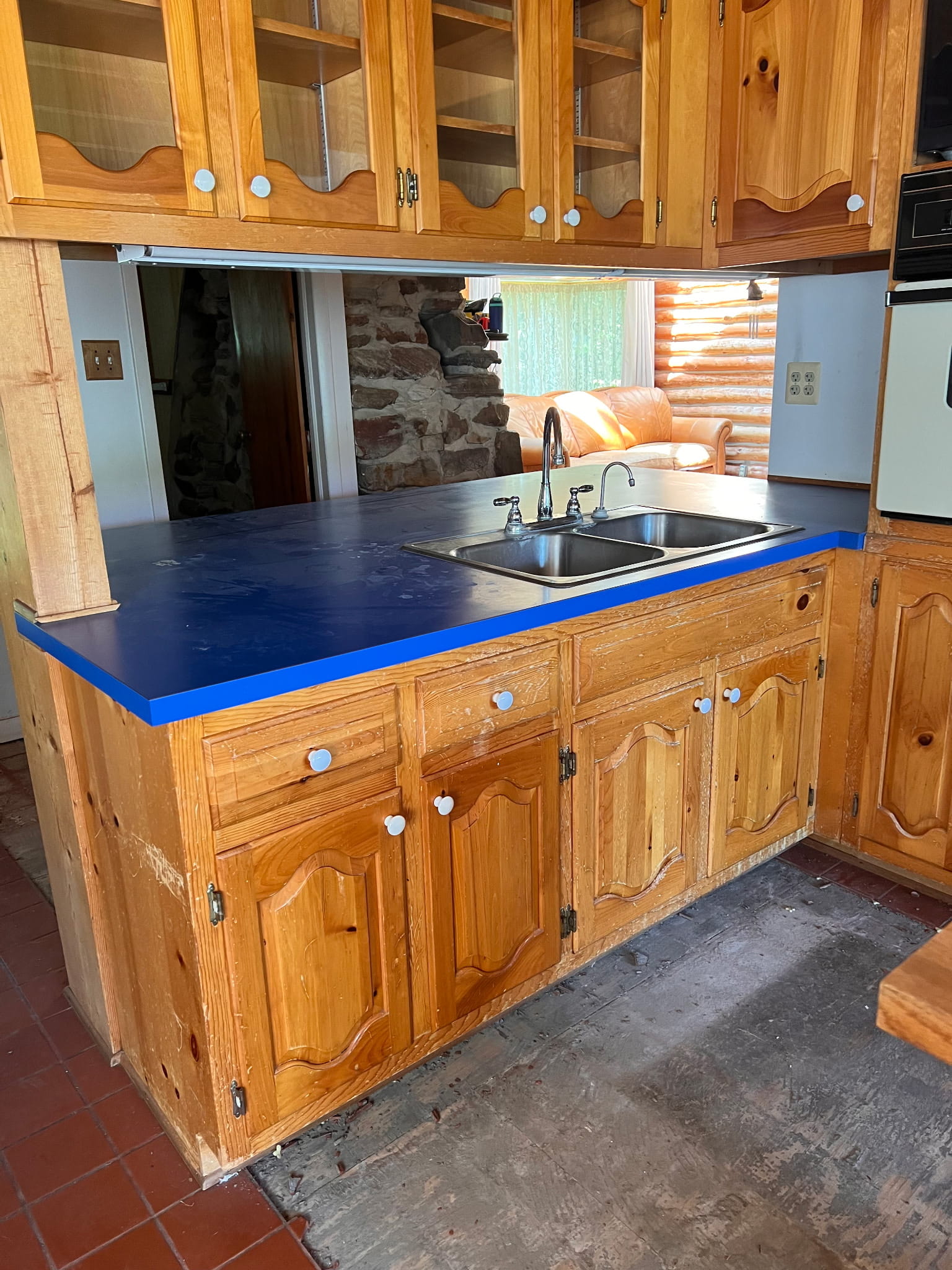 The original knotty-pine cabinets and cobalt-blue laminate countertop with a drop-in sink, looking through the pass-through to the log walls and stone fireplace beyond