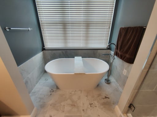 After: a freestanding white soaker tub under the window with a chrome floor-mounted tub filler, a deep teal wall, and marble-look tile