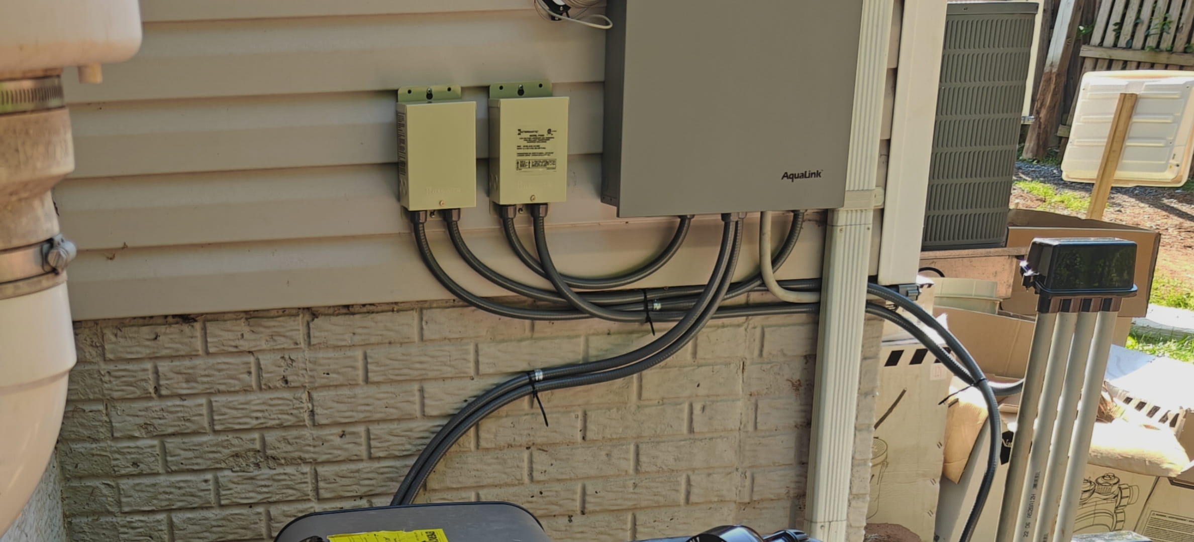 Close view of the Jandy AquaLink panel and two transformers mounted on the vinyl siding, with tidy flex conduit loops running down to each box