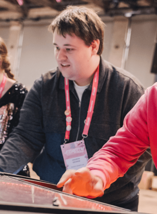 A man smiling and playing an arcade-style game at an event. He’s wearing a dark jacket over a white shirt with a pink lanyard around his neck. The background is softly blurred, showing other people and event lighting.