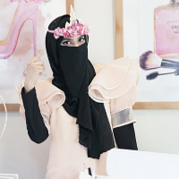 A woman wearing a black niqab and peach ruffled top stands in a bright crafting studio, smiling with her eyes and pointing upward while wearing a pink floral unicorn headband, with Cricut equipment and feminine art prints in the background.