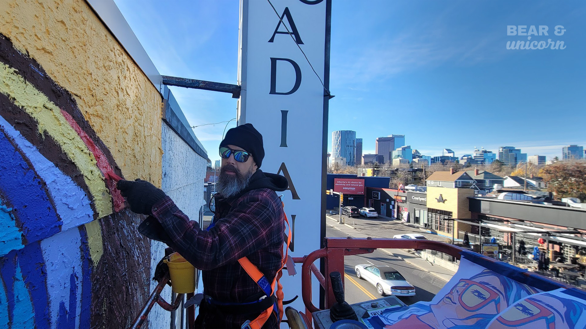 Artist David Schmeikal wearing a safety harness and sunglasses working from a boom lift to paint details on the high upper wall of the mural. Artist David Schmeikal wearing a safety harness and sunglasses working from a boom lift to paint details on the high upper wall of the mural.