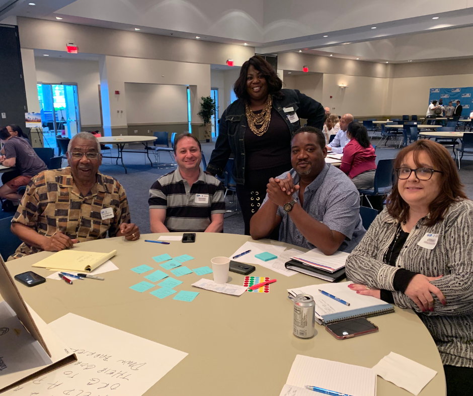A diverse group of executives and innovation leaders collaborate around a sleek boardroom table. Bianca Jaye Shelby leads an interactive AI strategy workshop, with digital screens displaying data and strategy visuals. The setting is bright, modern, and energetic, emphasizing teamwork and transformation.