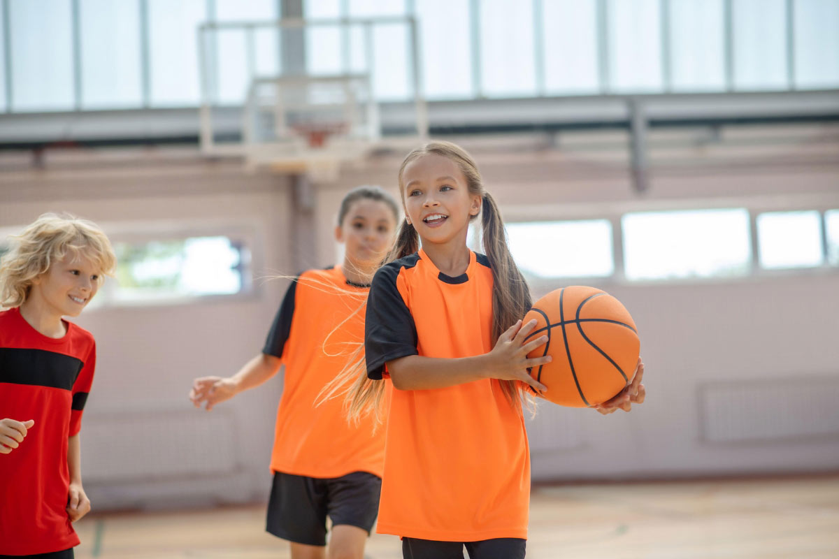 Children play basketball indoors, showing mobility and confidence while one child holds the ball and wears a prosthetic device.