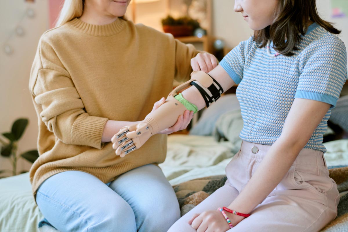 Child sits on a bed while a caregiver adjusts their prosthetic arm, ensuring proper fit and comfort.
