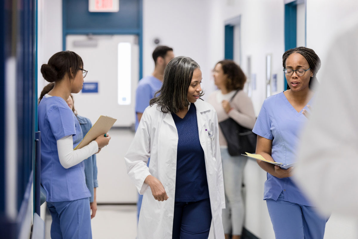 Diverse group of doctors, nurses, and hospital case managers in scrubs discussing medical files and walking down a busy hospital corridor. Diverse group of doctors, nurses, and hospital case managers in scrubs discussing medical files and walking down a busy hospital corridor.