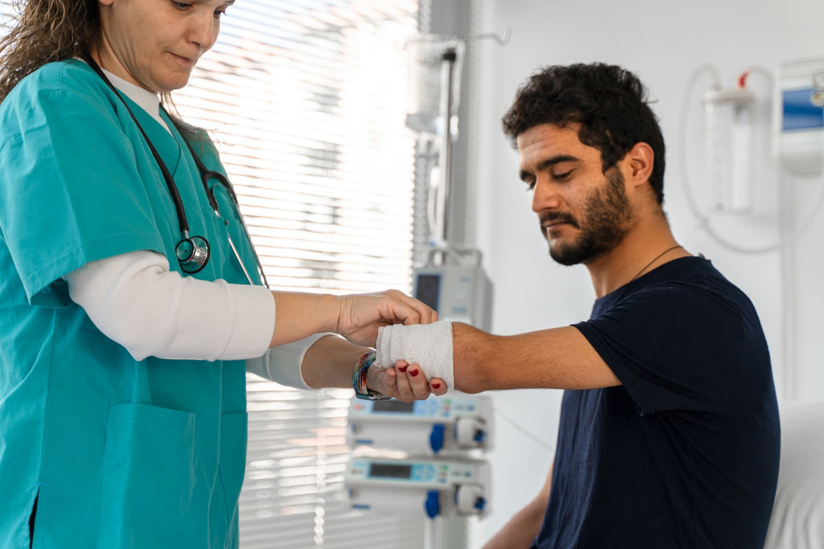 A female healthcare professional in teal scrubs carefully wrapping a compression bandage around a male patient's upper-extremity residual limb in a clinical hospital room. A female healthcare professional in teal scrubs carefully wrapping a compression bandage around a male patient's upper-extremity residual limb in a clinical hospital room.