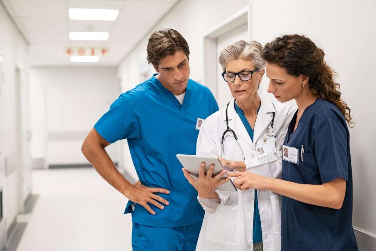 A group of three medical professionals in scrubs and lab coats discussing clinical data on a digital tablet in a bright hospital hallway. A group of three medical professionals in scrubs and lab coats discussing clinical data on a digital tablet in a bright hospital hallway.