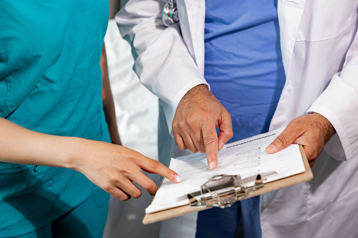 Close-up of a doctor in a white lab coat and a practitioner in teal scrubs reviewing clinical paperwork on a clipboard. Close-up of a doctor in a white lab coat and a practitioner in teal scrubs reviewing clinical paperwork on a clipboard.