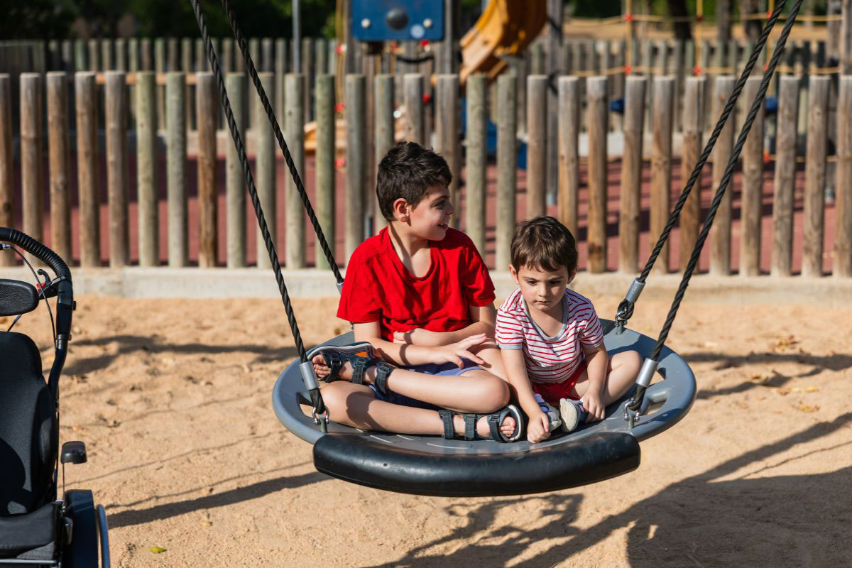 Two young boys sitting together on a large circular web swing at a sunny park playground