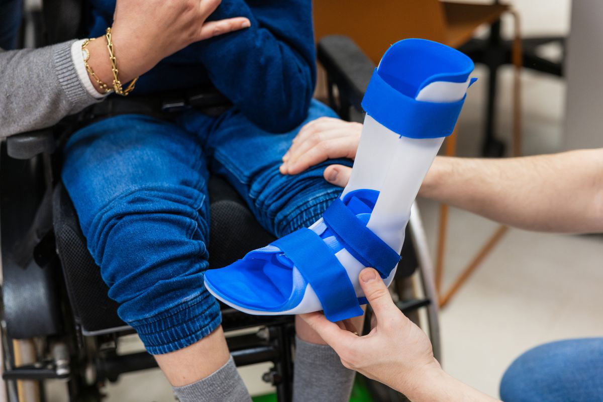 Hands of an orthotist adjusting a custom white and blue ankle-foot orthosis brace on a child sitting in a wheelchair